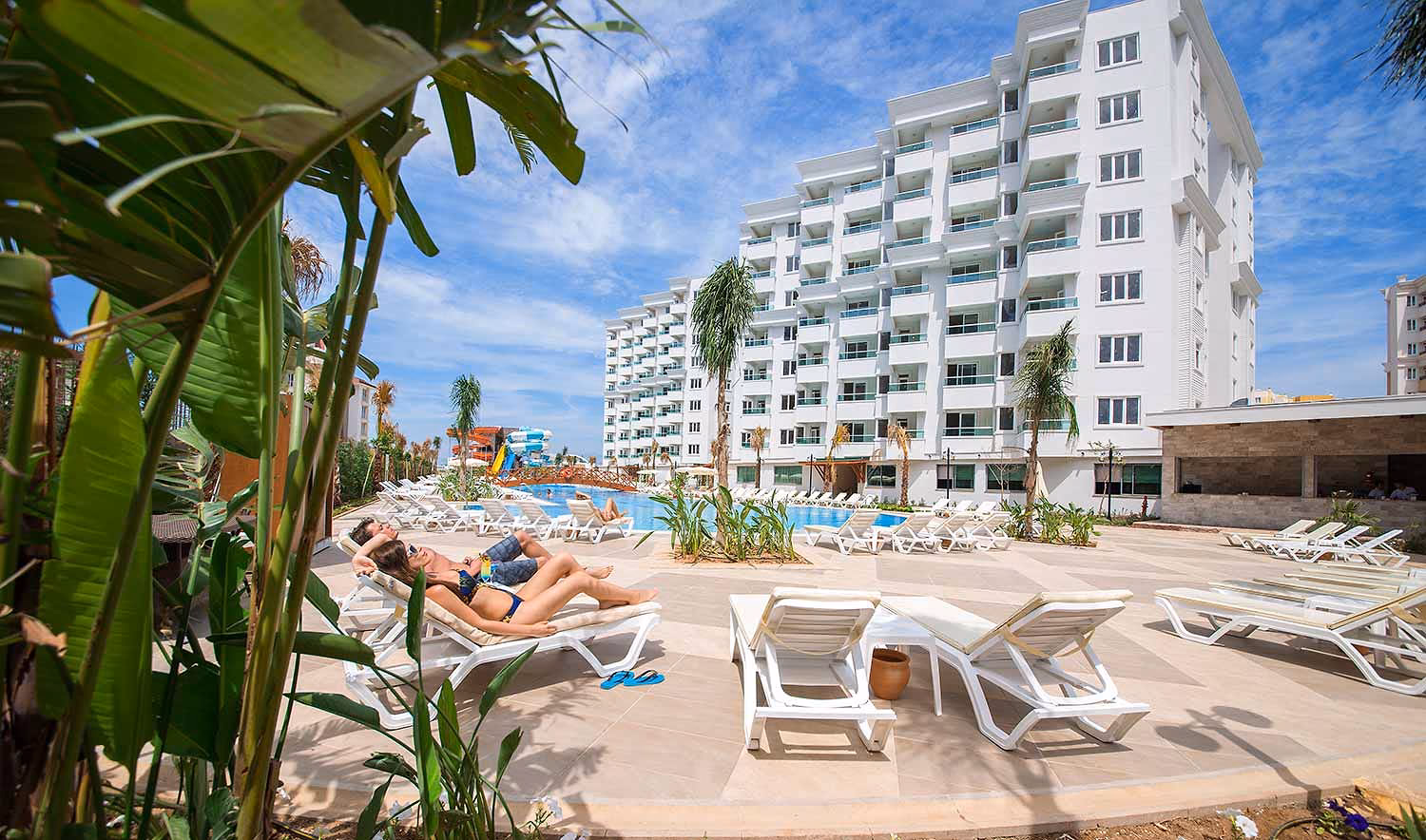 Twee zonnenden op ligbedden bij het zwembad, met de witte hotelgebouwen en op de achtergrond de glijbanen bij Wyndham Garden Lara in Lara bij Antalya