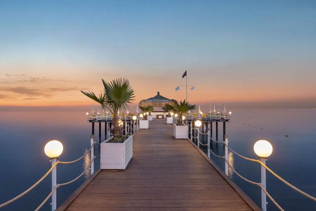 Houten pier met lampen en palmen in plantenbakken richting een rieten paviljoen aan zee bij zonsondergang bij Wyndham Garden Lara in Lara