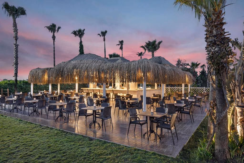 Overdekt openluchtterras met rieten daken, tafels en stoelen en een bar in de avondschemering bij Wyndham Garden Lara in Lara bij Antalya